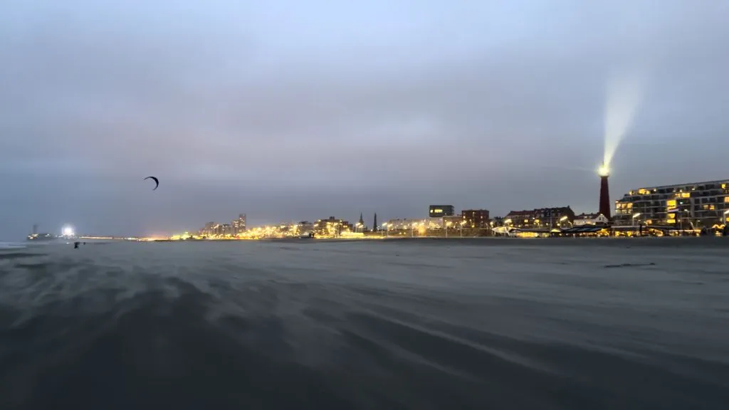 The Scheveningen beach on a windy evening. Blown sand hovers like a veil over the beach. The lighthouse casts its spotlight above all the other lighted buildings. In the distance the circular shape of the ferris wheel is glistening.